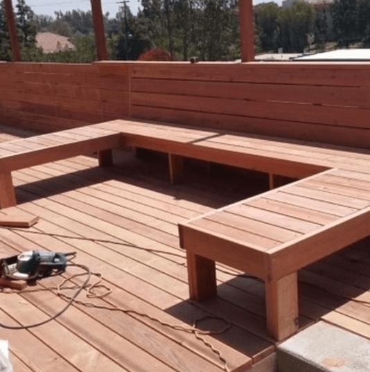 U-shaped wooden bench built into a redwood deck under construction with a power saw.